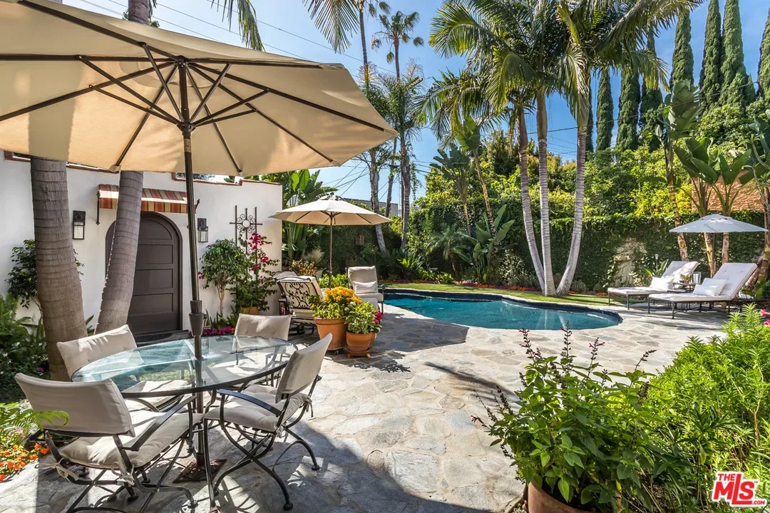 A backyard features a kidney-shaped pool, lounge chairs, umbrellas, and patio tables with chairs, surrounded by lush greenery and tall palm trees. A white stucco wall with a wooden door is in the background.